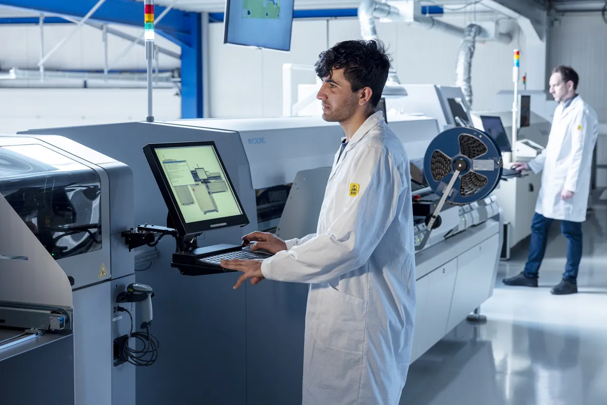 Electronics production in Belgium: a technician in an ESD coat at the screen of a pick-and-place machine, a colleague at the next machine.