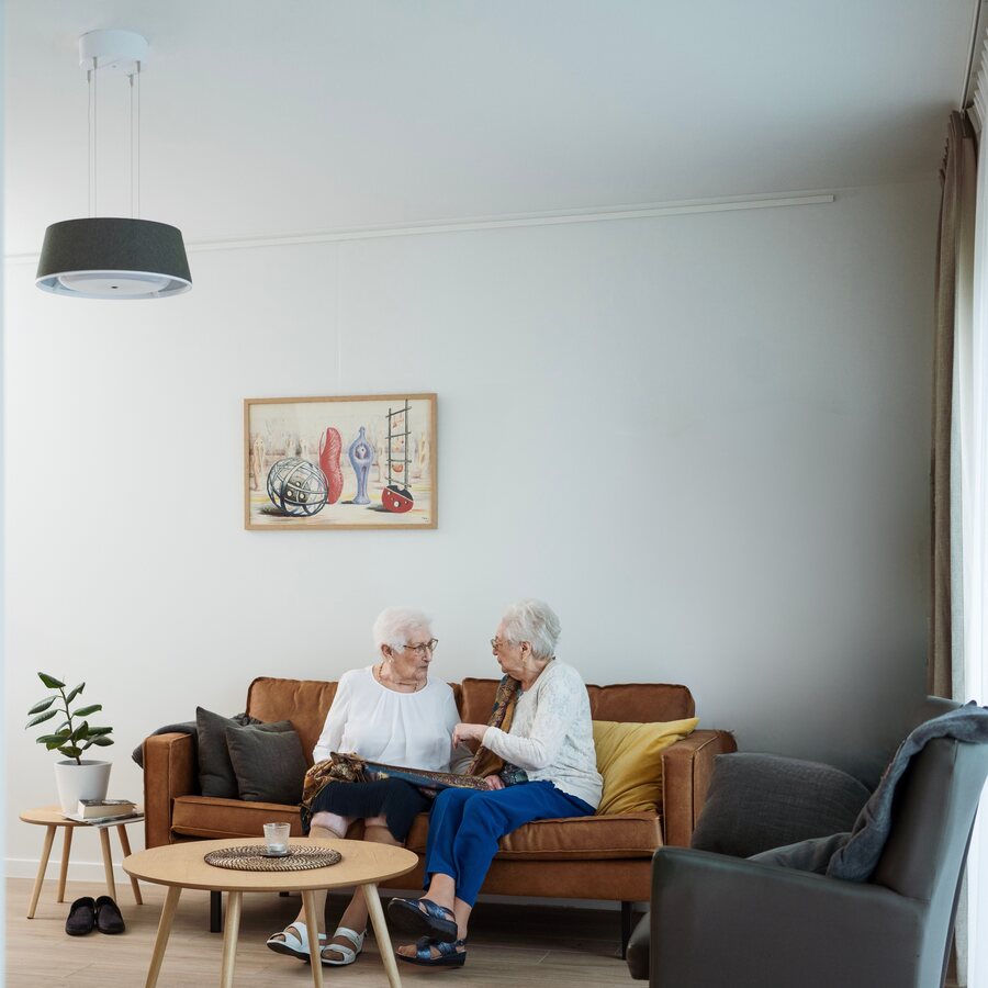 Two residents talking on the sofa in their apartment, under a Nobi Pendant light