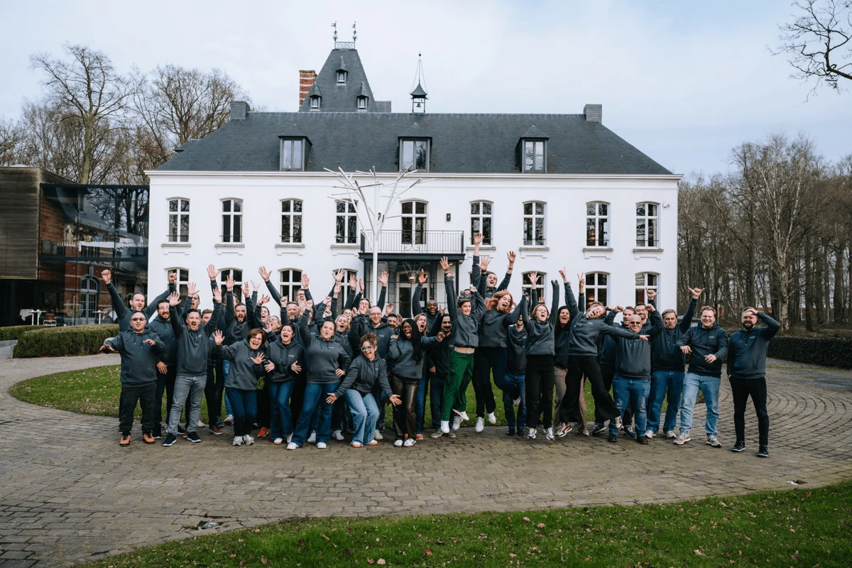 The Nobi team, more than fifty colleagues in grey Nobi hoodies with their arms in the air, in front of a white manor house.